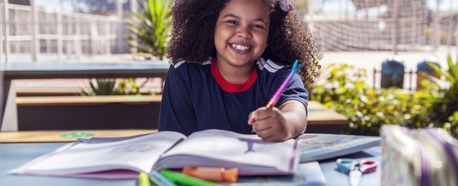 Uma criança feliz usando uniforme escolar, escrevendo com uma caneta de sereia em um livro, mostrando que o retorno à rotina escolar deve ser um momento feliz